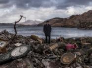 John Akomfrah, Video still from Vertigo Sea, 2015. A man in old timey garb stands on the shore of a pond littered with clocks.
