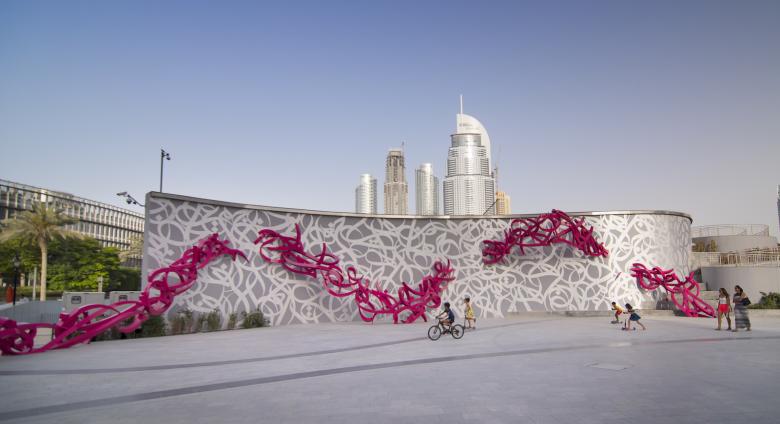 eL Seed, Declaration. Courtesy of the artist. a large installation piece in context—with city visible in the distance and children riding on bikes in front. 